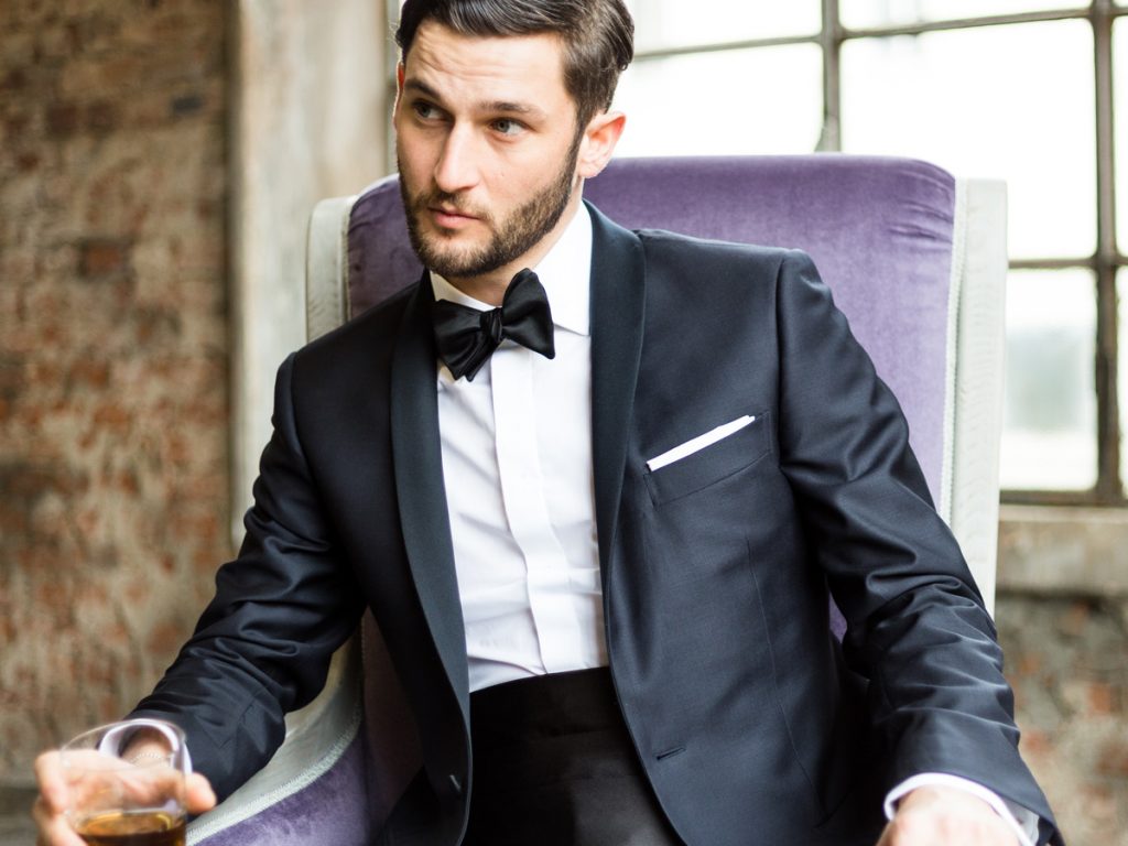Uomo seduto su una poltrona viola, indossa uno smoking nero di giorno con camicia bianca e papillon nero