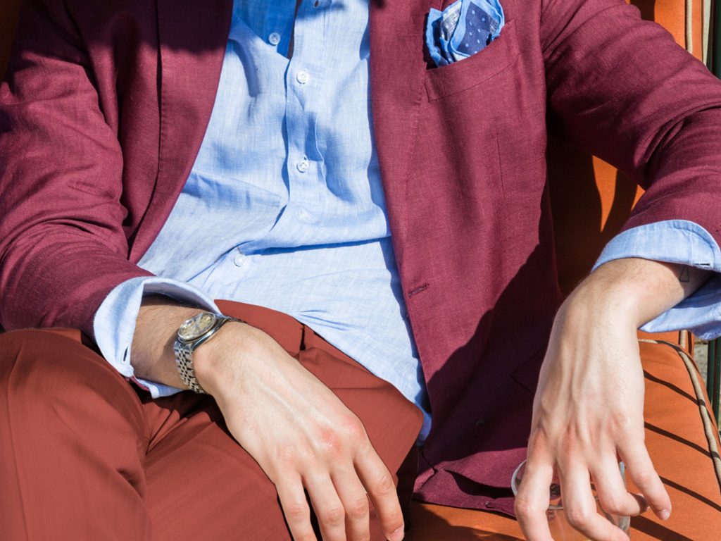 Dettaglio su un uomo che indossa uno spezzato tono su tono, giacca granata, camicia azzurra, pantaloni mattone e pochette azzurra