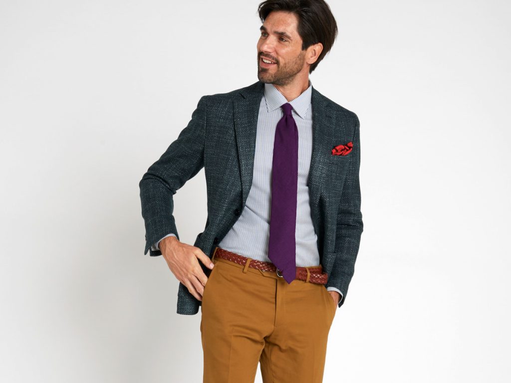 Un uomo indossa uno spezzato: una giacca petrolio, una cravatta viola, una camicia azzurra una pochette rossa, pantaloni ocra.